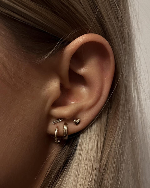 Mini huggie earring in 14k yellow gold, available as a single earring or pair. Glossy polished surface with a size of 11x1.6 mm. Model shown wearing multiple earring styles for earparty inspiration.